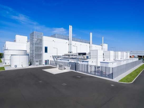 Das Bild zeigt ein modernes IndustriegebĂ€ude mit mehreren groĂen weiĂen Strukturen und Schornsteinen. Im Vordergrund befindet sich eine weitlĂ€ufige asphaltierte FlĂ€che. An der Seite des GebĂ€udes sind technische Anlagen wie Tanks und Rohrleitungen sichtbar. Die Architektur wirkt klar und sauber, und der Hintergrund zeigt einen blauen Himmel, was dem Bild eine helle und aufgerĂ€umte AtmosphĂ€re verleiht.