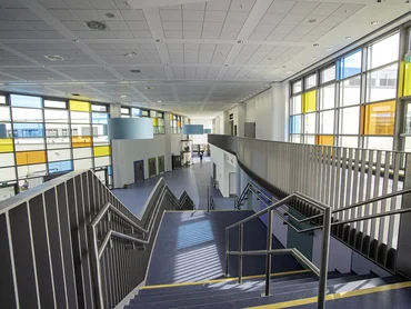 Foto: breiter Stiegenabgang mit Geländer aus Aluminium in langgezogenen Schulkorridor mit bunten Fenstern auf beiden Seiten, Decken mit integrierter Beleuchtung