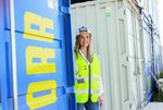 Picture shows a site manager standing next to a PORR construction container. She is wearing a helmet and a high-visibility waistcoat. The picture is an image image and symbolises PORR as a "modern employer"