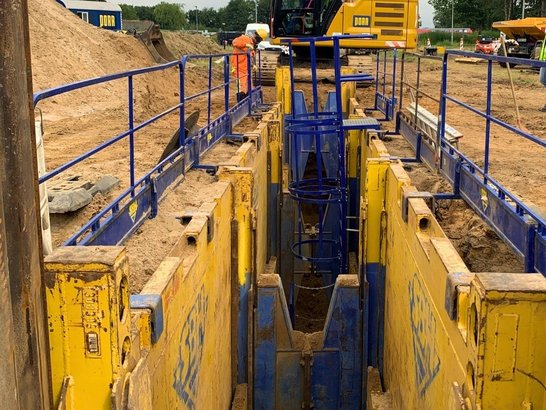 Baustelle im Leitungsbau mit gesichertem Graben, Bagger und Bauarbeitern in Schutzkleidung; umgeben von BĂ€umen und Baumaschinen.