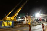A construction site at night on a road. A yellow crane lifts a steel part onto a concrete pillar.