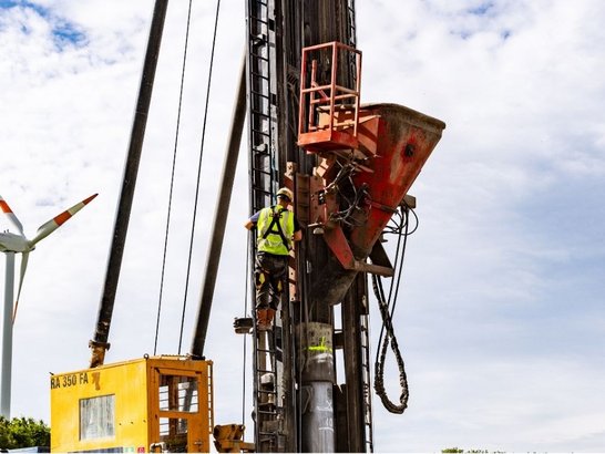 Ein Bauarbeiter in Sicherheitskleidung bedient eine groĂe Bohrmaschine mit gelber Kabine und orange-rotem Aufsatz auf einer Baustelle. Im Hintergrund steht ein Windrad mit orangefarbenen Rotorblattspitzen unter einem teils bewölkten Himmel.