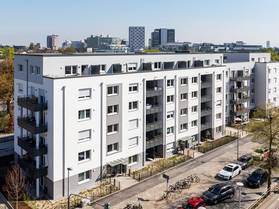 Mehrstöckiges WohngebĂ€ude mit weiĂ-grauer Fassade und Balkonen in urbaner Umgebung, mit geparkten Autos, FahrrĂ€dern und begrĂŒntem StraĂenbereich â Beispiel fĂŒr moderne Stadtarchitektur.