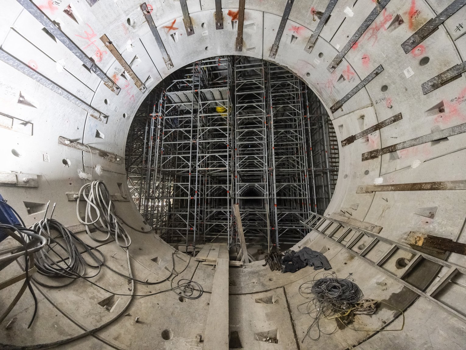 Innenansicht eines zylindrischen Bauwerks mit BetonwĂ€nden und umfangreichem GerĂŒstsystem, Kabeln und Werkzeugen â zeigt den Fortschritt bei einem Tunnel- oder Infrastrukturprojekt.