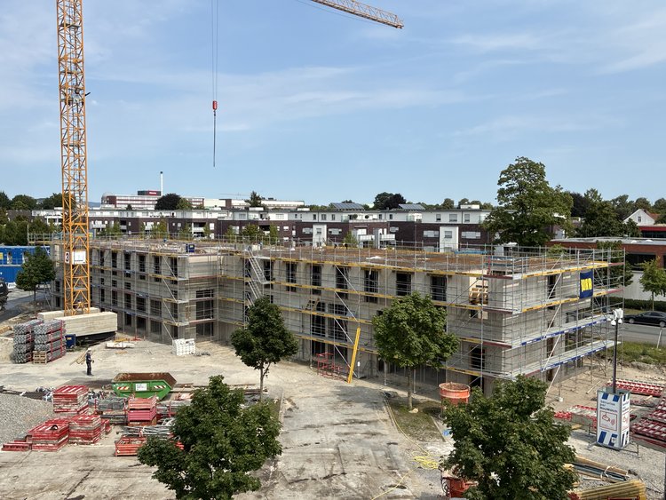 Das Bild zeigt eine Baustelle, auf der ein GebĂ€ude im Bau ist. Das GebĂ€ude ist teilweise fertiggestellt und von GerĂŒsten umgeben. Auf dem GelĂ€nde sind verschiedene Baumaterialien verteilt. Links im Bild befindet sich ein groĂer gelber Kran. Im Vordergrund sind mehrere BĂ€ume zu sehen, wĂ€hrend im Hintergrund WohngebĂ€ude und ein klarer blauer Himmel erkennbar sind.
