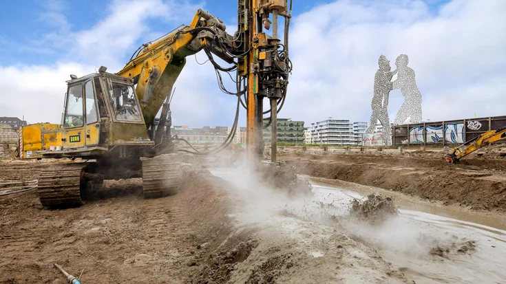 Bild zeigt ein Bohrgerät beim Herstellen einer Düsenstrahlsohle in Berlin; Im Hintergrudn sieht man das bekannte Kunstwerk Berlin und im Vordergrund spritzt der Dreck vom Herstellungsprozess der Dichtsohle. Das Düsenstrahlverfahren in einem Bild