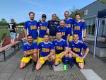 9 men in blue and yellow jerseys stand together for a group photo. In the background you can see a sports field.