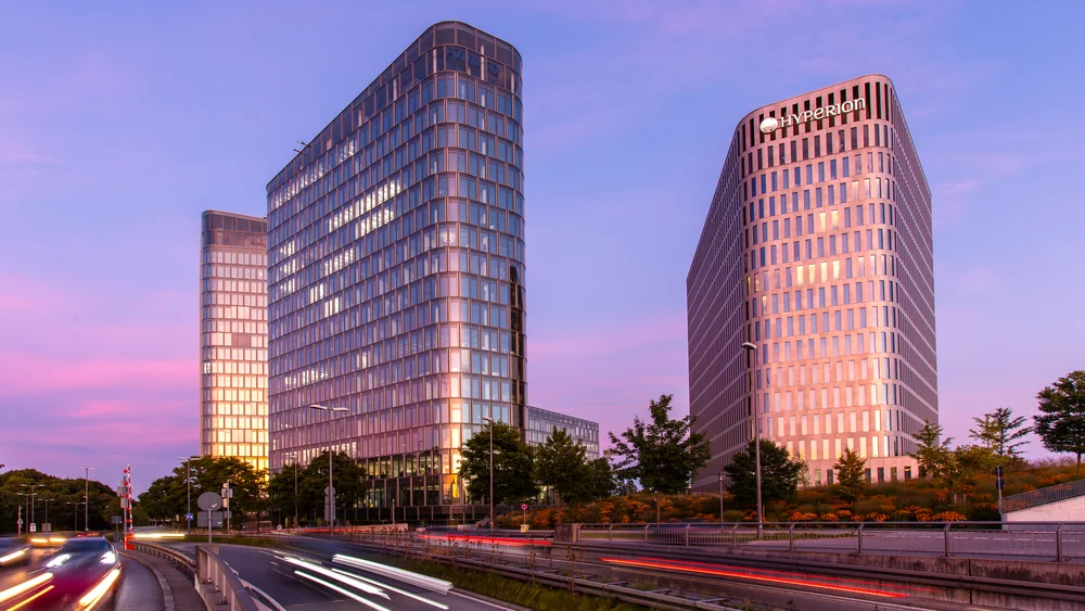 View of the two high-rise buildings of the hotel tower; on the left you can see the Blue Tower which is an office building; on the right you can see a hotel belonging to the Hyperion brand; the floor-to-ceiling glazing of the windows is striking; picture at dusk.