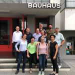A group of men and women stand together on a staircase and laugh into the camera. The words Bauhaus can be seen on the building behind them.