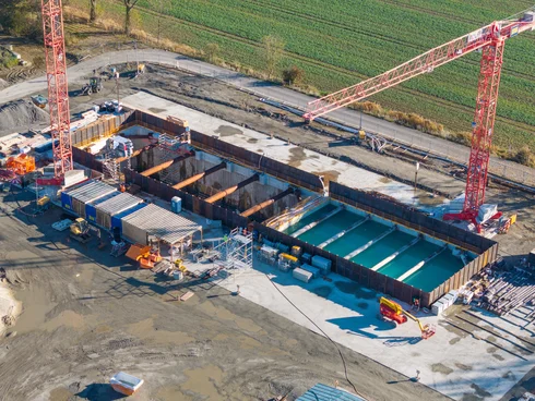 Bild zeigt Baugrube von der Seite. Man sieht das die rechte Seite mit blauen Wasser voll ist. Drum herum stehen Baumaschinen und Kräne