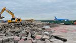  View of a broken up tarmac surface. A blue aeroplane and an excavator can be seen in the background.