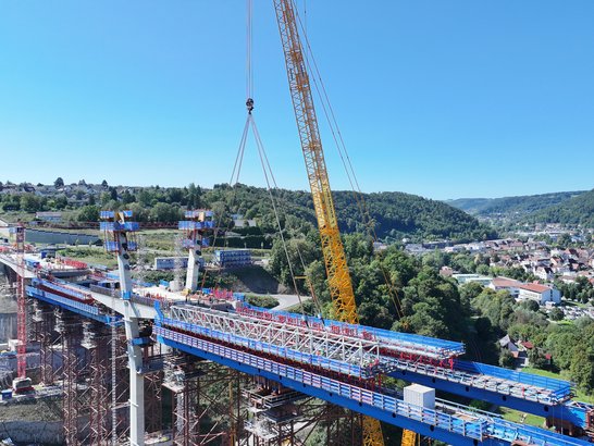 GroĂbaustelle fĂŒr eine BrĂŒcke: Ein gelber Kran hebt ein groĂes Bauteil ĂŒber ein Tal, wĂ€hrend Arbeiter auf der BrĂŒcke arbeiten. Im Hintergrund sind grĂŒne HĂŒgel, eine Stadt und blaue GerĂŒste sichtbar.