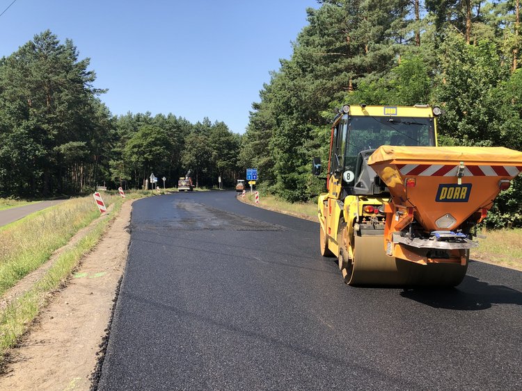 Kreisverkehrsumbau und Fahrbahnsanierung, L 23 / L 38 Das Bild zeigt eine StraĂenszene wĂ€hrend Bauarbeiten. Ein gelbes StraĂenwalzenfahrzeug mit der Aufschrift "PORR" legt eine neue Asphaltschicht auf eine lĂ€ndliche StraĂe, die von BĂ€umen und GrĂŒnflĂ€chen umgeben ist.