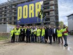 A few people are standing in front of a building shell, on which hangs a large poster with the inscription "PORR" in yellow. A topping-out wreath hanging from a crane can be seen above the people. 
